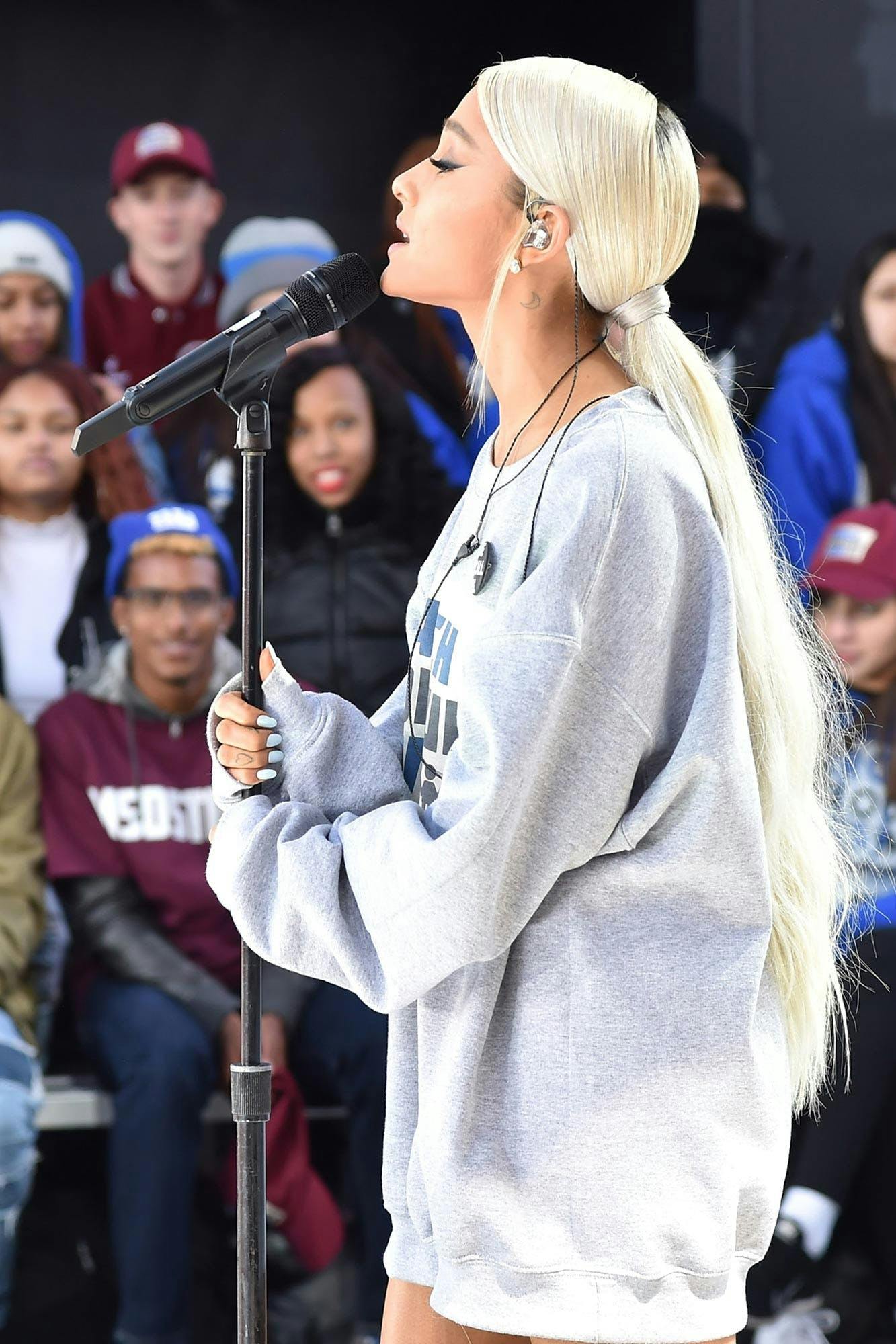Ariana Grande singing in a grey sweatshirt and a blonde low ponytail.