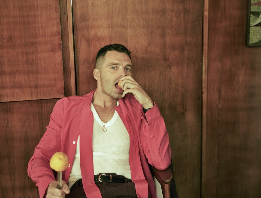Sebastian stan sits in a chair with his legs open in brown trousers, a white tank top, an open pink shirt, and an orange on a knife in one hand and an orange in his mouth in the other.