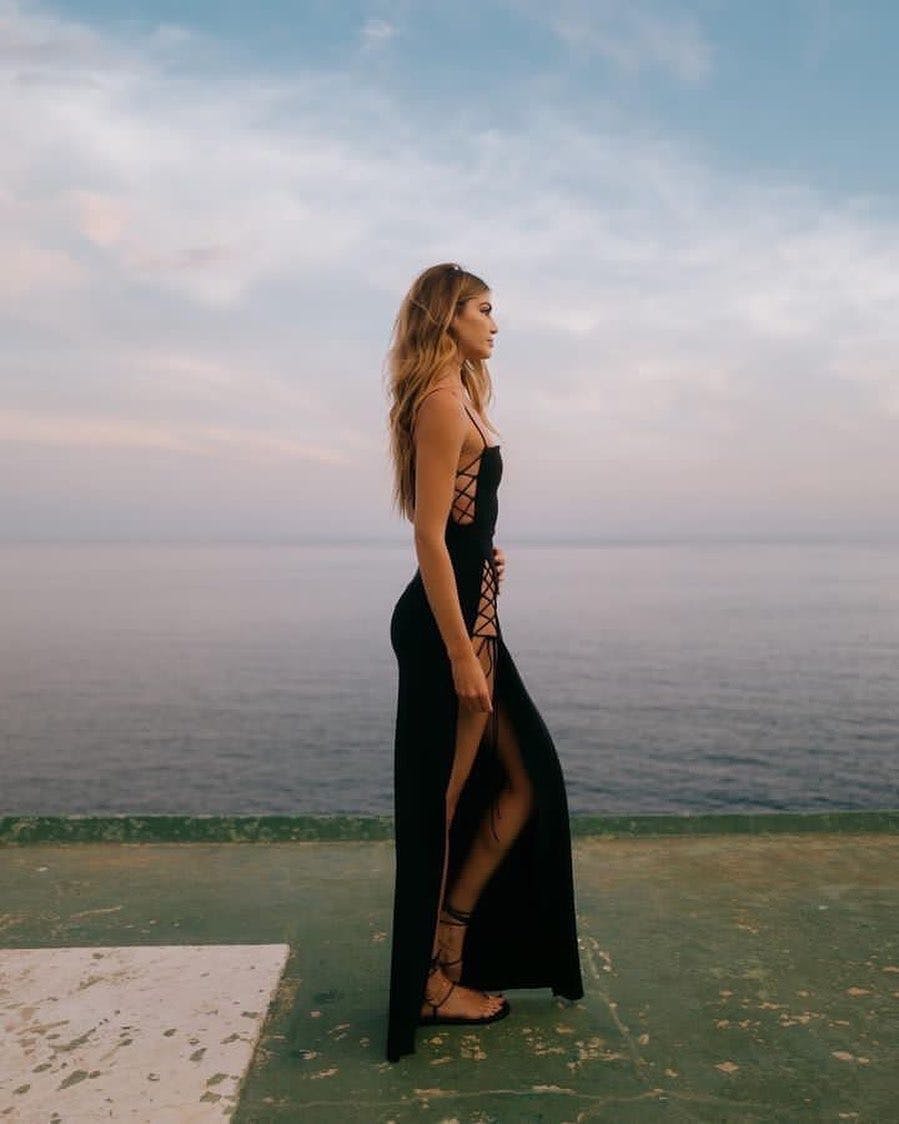 Model wearing a black dress with a side slit while standing in front of the water.